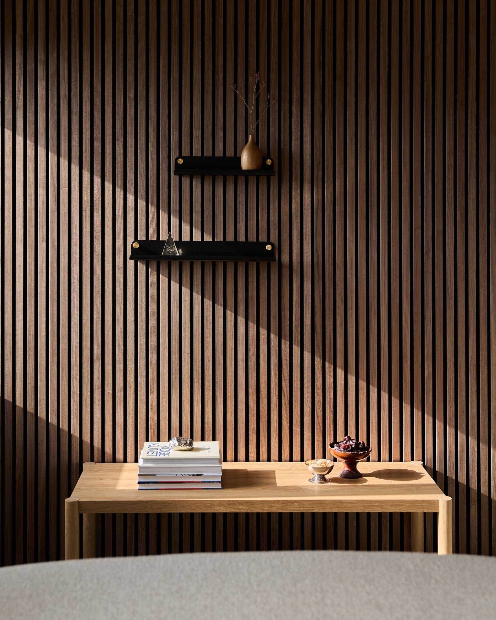 Ett minimalistiskt träbord med böcker och skålar står intill en nordisk ribbvägg Akustikpanel och lyfter fram WoodUpp Norr Narrow Shelf i black (51,9 cm), med solbelysta skuggor ovanför.